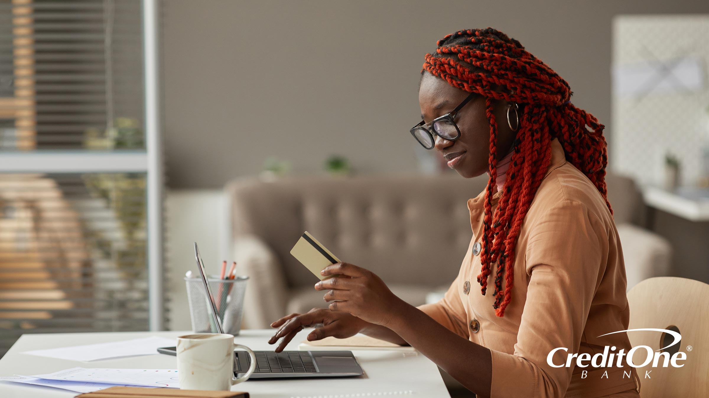A young woman uses her laptop while looking at her credit card as if she’s shopping online. Perhaps she’s making use of her credit card’s grace period.